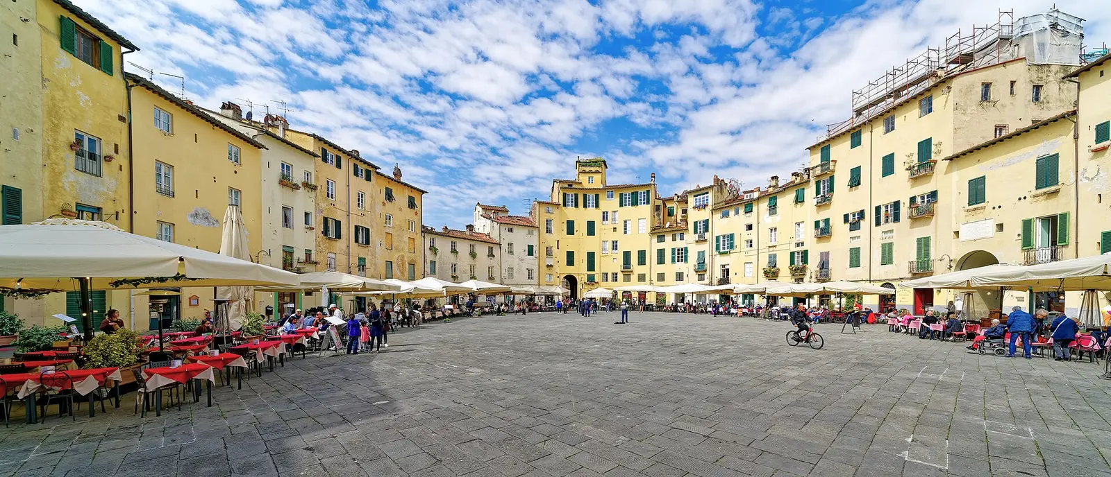 The oval Piazza dell'Anfiteatro in Lucca with its ring of ochre houses and cafe tables under a cloudy blue sky